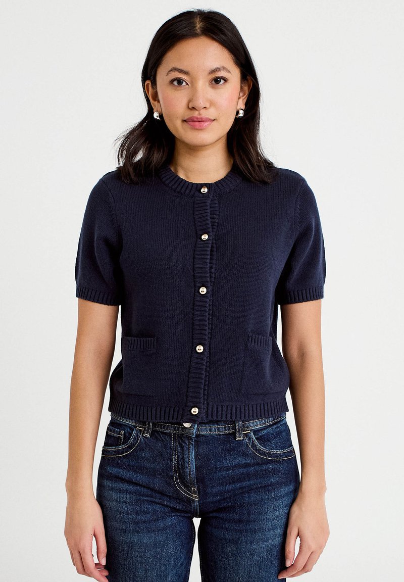 Frau trägt eine marineblaue, kurzärmelige, mit Knöpfen versehene Strickjacke mit zwei aufgesetzten Taschen vorne und dunkelblaue Jeans, steht vor einem schlichten Hintergrund.