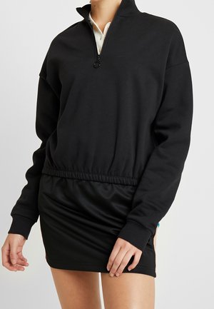 Svart tröja med halv dragkedja, elastisk midja och långa ärmar. Mjuk material med avslappnad passform; har en sidofliksdetalj.
