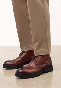 Stivali brogue in pelle marrone con una finitura liscia, dotati di perforazioni decorative, suola in gomma nera e lacci a contrasto, abbinati a pantaloni khaki.