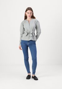 Sivý peplum top s previazaním v páse, dlhými rukávmi a hlbokým V výstrihom, skombinovaný s modrými úzkymi džínsami a čiernymi oxfordkami.