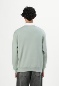 Persoon met kort, krullend donker haar, gekleed in een lichtgroene sweatshirt over een wit shirt en donkergrijze spijkerbroek, met de rug naar de camera op een effen witte achtergrond.