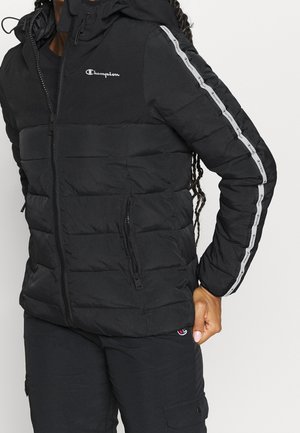Person wearing black Champion puffer jacket with logo on chest and branded stripes along sleeves, paired with black pants.