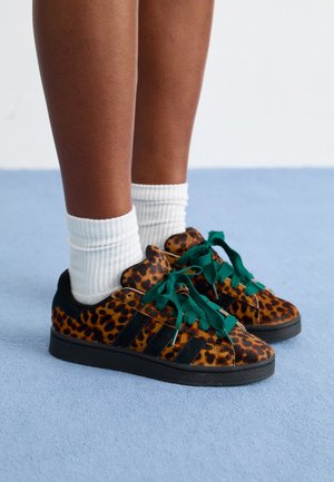 Beine mit dunkler Haut, die weiße gerippte Socken und Leoparden-Sneakers mit schwarzen Streifen und grünen Schnürsenkeln tragen, stehen auf blauem Teppich.