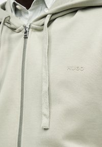 Ανοιχτό γκρι φούτερ με φερμουάρ και κορδόνια, φορεμένο πάνω από ένα λευκό πουκάμισο με γιακά, με ανάγλυφο λογότυπο "HUGO" στο στήθος.