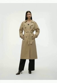 Trench coat bege com botões duplos, cintura cinto, bolsos laterais e mangas largas, combinado com calças pretas e saltos altos.