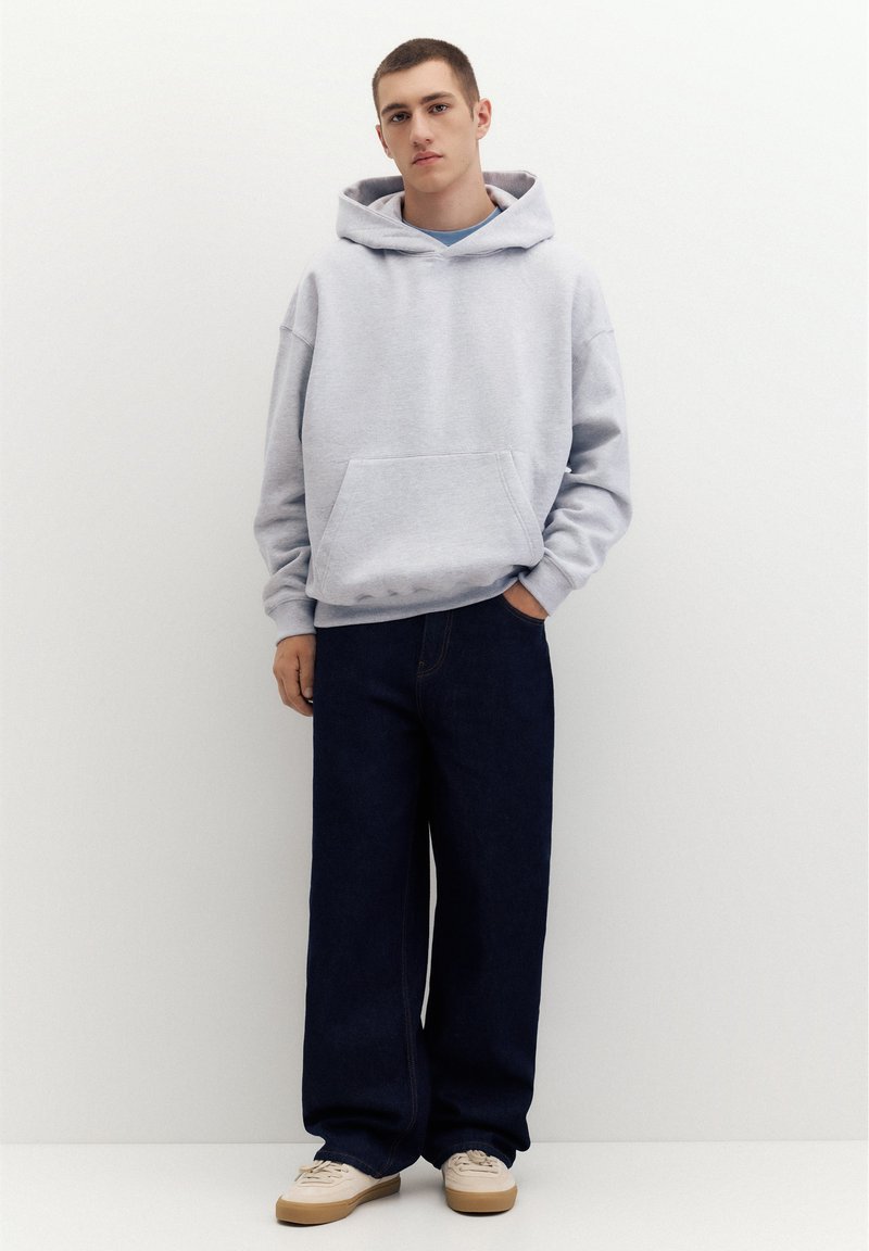 Jeune homme portant un sweat à capuche gris clair, un jean large foncé et des baskets beiges, debout devant un fond blanc uni.