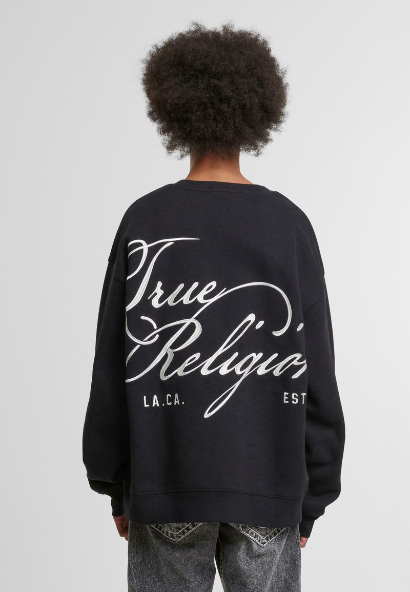 Sweatshirt noir à coupe oversized, avec un texte scripté blanc "True Religion" au dos. Poignets et ourlet côtelés, matériau en coton doux.