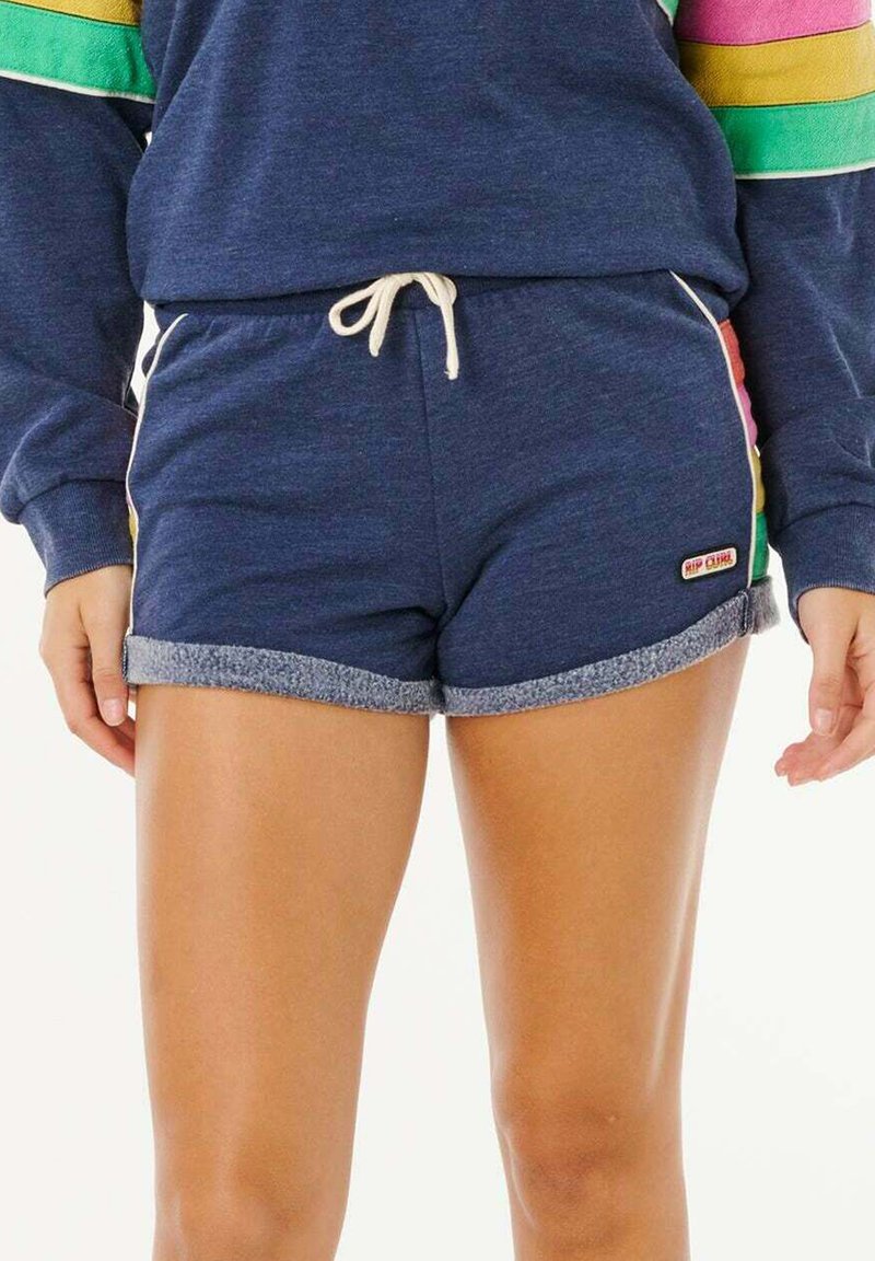 Person mit marineblauen Shorts mit umgeschlagenen Saumkanten und weißem Kordelzug, kombiniert mit einem langärmligen Oberteil mit bunten Streifen an den Ärmeln.