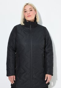 Kvinna med rakt blont hår som bär en svart vadderad jacka med dragkedjefickor och hög krage mot en enfärgad vit bakgrund.