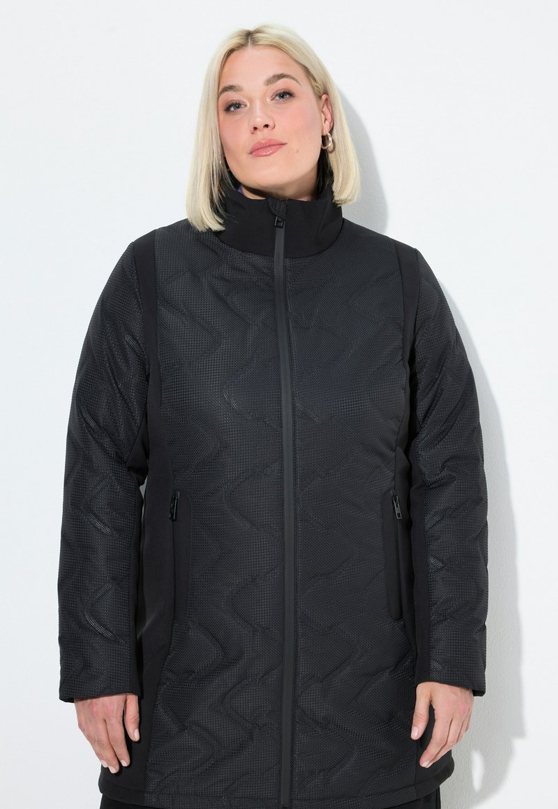 Kvinna med rakt blont hår som bär en svart vadderad jacka med dragkedjefickor och hög krage mot en enfärgad vit bakgrund.