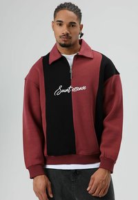 Sweat-shirt zippé bordeaux et noir avec col, présentant un logo blanc à l'avant, confectionné dans un tissu doux et texturé.