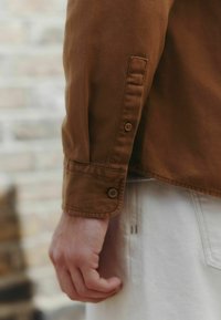 Hnedá košeľa na gombíky s manžetou a dvoma gombíkmi, vyrobená z textúrovanej tkaniny; ruka položená na svetlom oblečení.