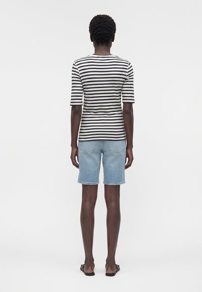 Άτομο που στέκεται με την πλάτη στραμμένη, φοράει μαύρο-άσπρο ριγοειδές πουκάμισο με μανίκια μέχρι τους αγκώνες, ανοιχτά τζιν σορτς και μαύρα σανδάλια.