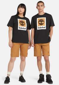 Svarte bomulls t-skjorter med Timberland-logo, kombinert med beige shorts. Begge har på seg svart fottøy og stripete sokker.