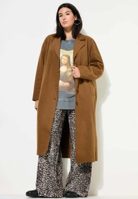 Bruine oversized jas met zijsplitsen, gedragen over een grafisch sweatshirt en een pant met luipaardprint met wijde pijpen. Zwarte enkellaarsjes maken de outfit compleet.
