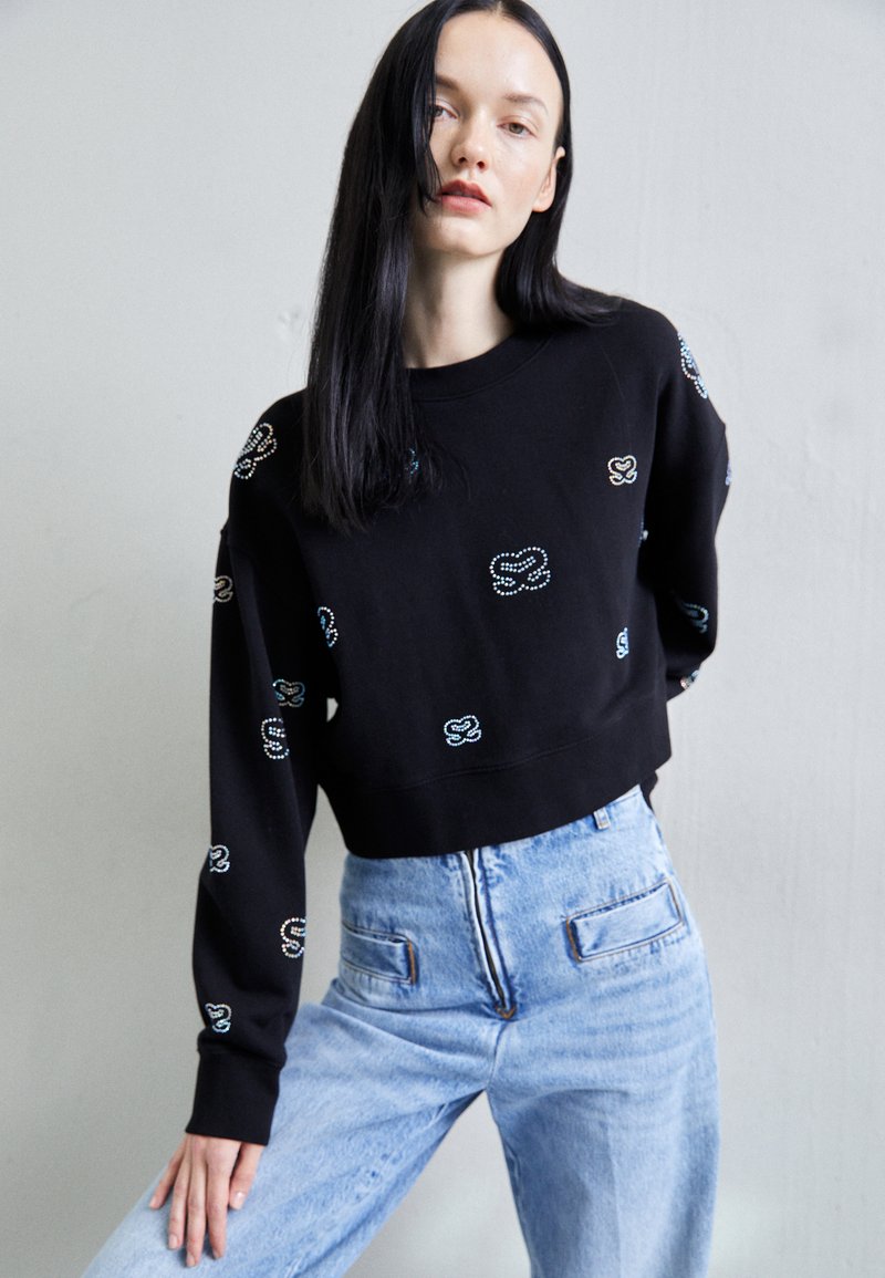 Schwarzer, kurz geschnittener Sweatshirt mit buntem besticktem Design, runder Halslinie und langen Ärmeln, kombiniert mit hoch geschnittenen hellblauen Jeans.