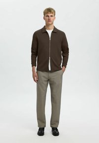 Maglione marrone con zip su una camicia bianca, abbinato a pantaloni grigi chiari e scarpe nere. Il tessuto appare liscio con una vestibilità strutturata.