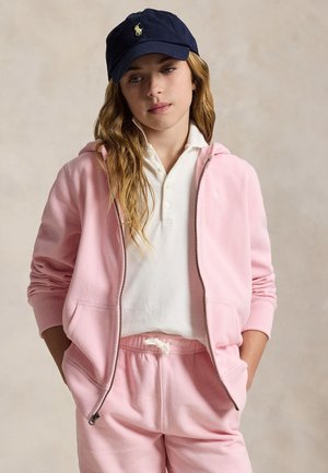 Mladá žena s tmavomodrou čiapkou, bielym polokošľom a ružovou mikinou na zips so zodpovedajúcimi ružovými teplákmi, stojaca s rukami v kapsách.