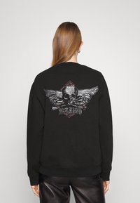 Felpa nera con design di teschio decorato in argento e rosso con ali sul retro. Polsini e orlo a costine, tessuto morbido.
