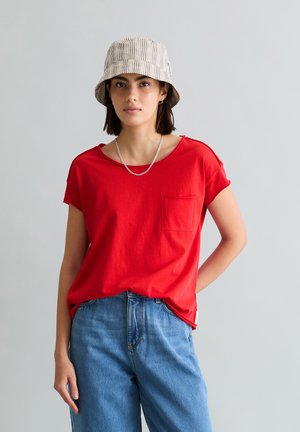 Junge Frau mit einem beigen, karierten Anglerhut, rotem T-Shirt mit Taschen, silberner Gliederkette und blauer Jeans vor einem einfachen grauen Hintergrund.