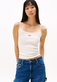 Débardeur blanc à côtes avec des bretelles fines, orné d'un logo rouge "tommy jeans" au centre. Associé à un jean en denim bleu.