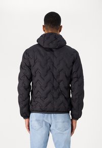 Giacca a vento nera trapuntata con cappuccio, caratterizzata da un motivo zigzag e polsini elastici, abbinata a jeans chiari strappati.