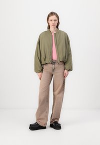 Světlý olivově zelený bomber, růžový top, hnědé široké kalhoty a černé platformové boty. Model stojí proti jednoduchému bílému pozadí.