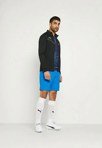 Άνδρας ντυμένος με μπλε σορτς, μαύρο μπουφάν, λευκές κάλτσες μέχρι το γόνατο και λευκά παπούτσια κατά της απλής ανοιχτόχρωμης φόντου.