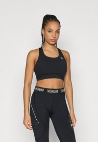 Melns sporta krūšturis ar noapaļotu kakla izliekumu un logotipu, kopā ar melnām legingām ar augstu jostasvietu, kurām ir zīmola jostasvieta un baltas detaļas.