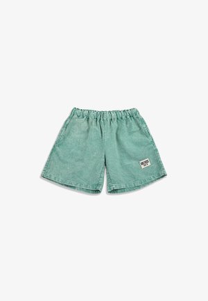 Zelené elastické krátke nohavice s malou bielou obdĺžnikovou záplatou na ľavej nohe s nápisom "BRON CROSS SINCE 1991."