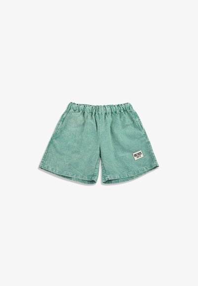 Shorts verts à taille élastique avec une petite étiquette rectangulaire blanche sur la jambe gauche indiquant "BRON CROSS SINCE 1991".