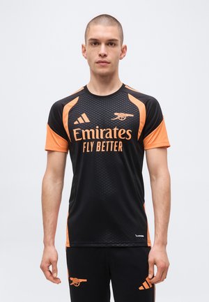 Giovane uomo che indossa la maglia da calcio nero e arancione dell'Arsenal con il logo Emirates Fly Better, in piedi davanti a uno sfondo chiaro e uniforme.