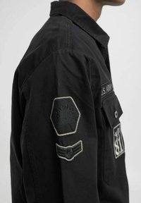Sort skjorte med militær-inspirerede lapper på venstre ærme, herunder hexagon- og vinge-designs. Enkel, holdbar stof med en afslappet pasform.