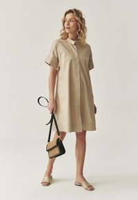 Beige lyhythihainen mekko, jossa on korkea kaulus, sileä pinta ja suora leikkaus. Yhdistettynä mustaan käsilaukkuun, jossa on kudottu etuosa, sekä litteisiin sandaaleihin.
