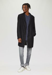 Schwarzer Mantel mit Reverskragen, Knopfverschluss und zwei Taschen, getragen über einem marineblauen Hemd mit Knöpfen und hellgrauen Jeans mit umgeschlagenen Säumen.