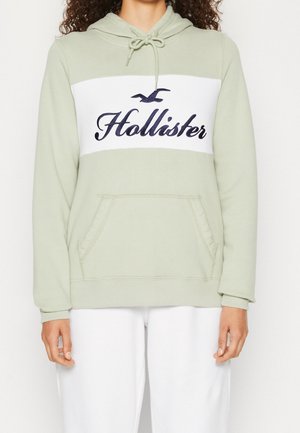 Persona con sudadera con capucha verde claro con panel blanco y logo "Hollister" en azul oscuro, combinada con pantalones blancos, de pie contra un fondo blanco.