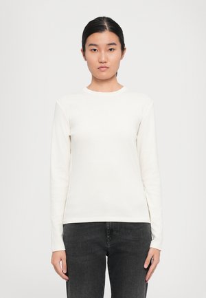 Young woman with dark hair tied back, wearing a white long-sleeve top and dark gray jeans, standing against a plain light background.
