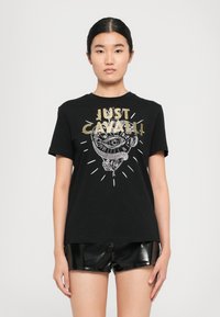 Young woman wearing black graphic "Just Cavalli" t-shirt with eye and snake design, paired with shiny black shorts, standing against white background.