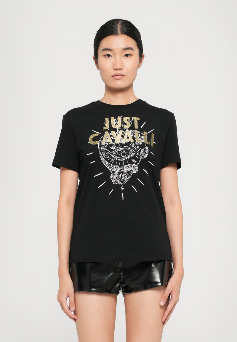 Young woman wearing black graphic "Just Cavalli" t-shirt with eye and snake design, paired with shiny black shorts, standing against white background.
