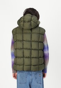Vesta căptușită de culoare verde măsliniu cu glugă, având un design matlasat și o suprafață texturată. Purtați peste un pulover multicolor și blugi.