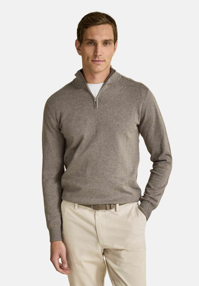 Homme portant un pull marron clair à zip quart, sur une chemise blanche avec un pantalon beige et une ceinture assortie, debout avec une main dans la poche.