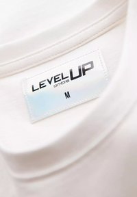 Bílý límec trička s lesklou visačkou s nápisem "LEVEL UP" a "ombre", velikost M, s hladkou strukturou a čistým švem.