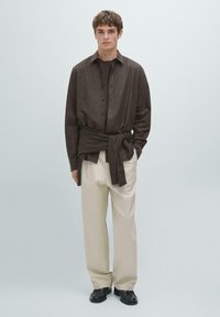 Brown button-up shirt made of soft fabric, paired with cream-colored wide-leg trousers. A dark sweater is tied around the waist. Black shoes.
