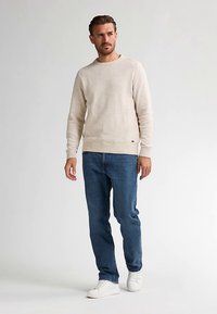 Beige gebreide trui met geribbelde manchetten en zoom, gecombineerd met een blauwe spijkerbroek en witte sneakers. Model staat nonchalant tegen een neutrale achtergrond.
