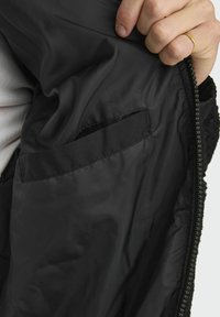 Interior de chaqueta negra con tejido suave, un pequeño bolsillo y una cremallera. La capa exterior presenta un material texturizado, posiblemente de forro polar.