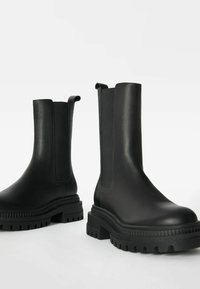 Botas de tobillo de cuero negro con paneles laterales elásticos, suela acanalada y punta redondeada. Textura suave con tiradores para facilitar el calzado.
