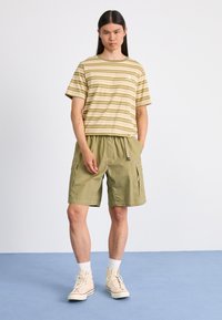 Camisa de manga corta a rayas en verde y beige, combinada con pantalones cortos de cargo en oliva. Zapatillas altas blancas y calcetines blancos completan el conjunto.