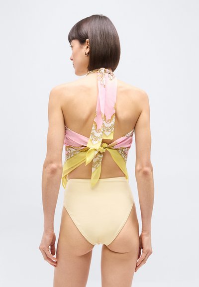 Γυναίκα με κοντά σκουρόχρωμα μαλλιά, που φοράει τοπ παστέλ ροζ και κίτρινο με δέσιμο στο πίσω μέρος και μπεζ σορτς ψηλόμεσα, στέκεται μπροστά σε ένα απλό φόντο.