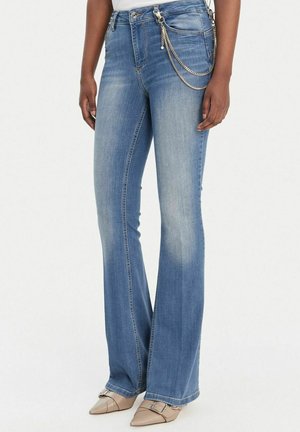 Jeans bootcut a vita alta blu con parte frontale slavata, accessorio a catena argento sulla tasca destra, indossati con scarpe beige a punta con fibbia.