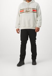 Felpa con cappuccio grigia chiara con tasca frontale a marsupio, caratterizzata da un design a righe marroni e rosse e dal logo Lacoste. Abbinata a pantaloni neri.